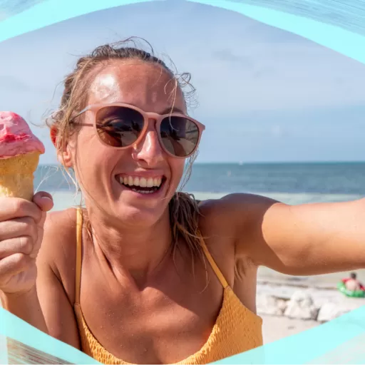 A young woman taking a selfie at the beach while holding a pink ice cream cone, wearing sunglasses, with the ocean in the background.