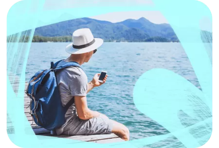 Man sitting on a wooden dock by a large lake, wearing a hat and backpack, and holding a smartphone with mountains visible in the background.