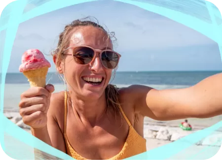 A young woman taking a selfie at the beach while holding a pink ice cream cone, wearing sunglasses, with the ocean in the background.